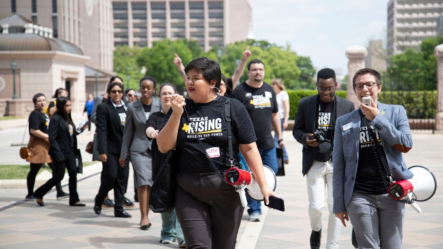 A group of protesters walk wearing Texas Freedom Network Education Fund shirts. The two people at the front speak into bullhorns
