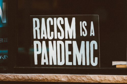 A sign in a window reads Racism is a Pandemic. Photo by Jon Tyson 1200x800