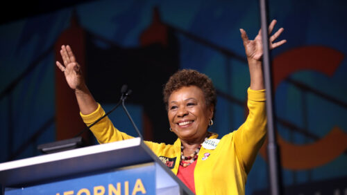 Photo of Representative Barbara Lee, a Black woman with short textured hair, standing on a podium and smiling and waving. She is wearing a yellow jacket and orange shirt