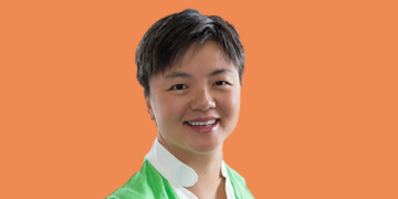 Photo of Sharon Chen, an asian woman with short grey hair smiling at the camera. She is wearing a green shirt with a white collar. The background of the photo is orange.