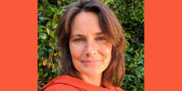A photo of WDN member Amanda Coslor, she has short brown hair, blue eyes, and is smiling slightly at the camera. She wears an orange sweater and is outside in front of a bush.