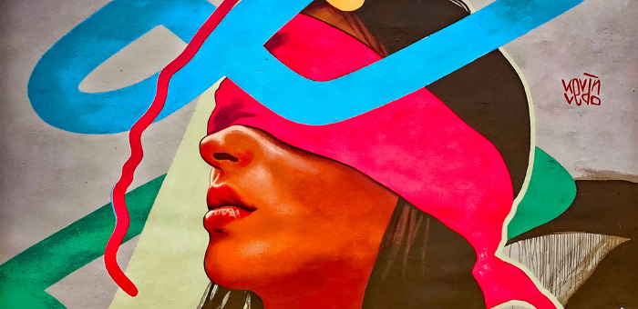 Photo of a colorful mural. The mural depicts a person with long hair with a bright pink blindfold tied over her eyes. Colorful lines and loops are drawn in the background, some cover part of the person's face