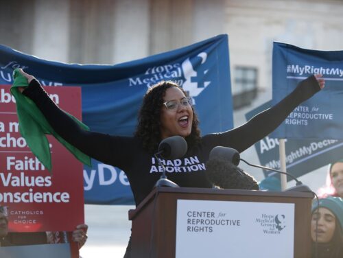 Photo of a person speaking at a podium in front of the supreme court of the US. Banners banners are behind them. Their shirt reads "I had an abortion."