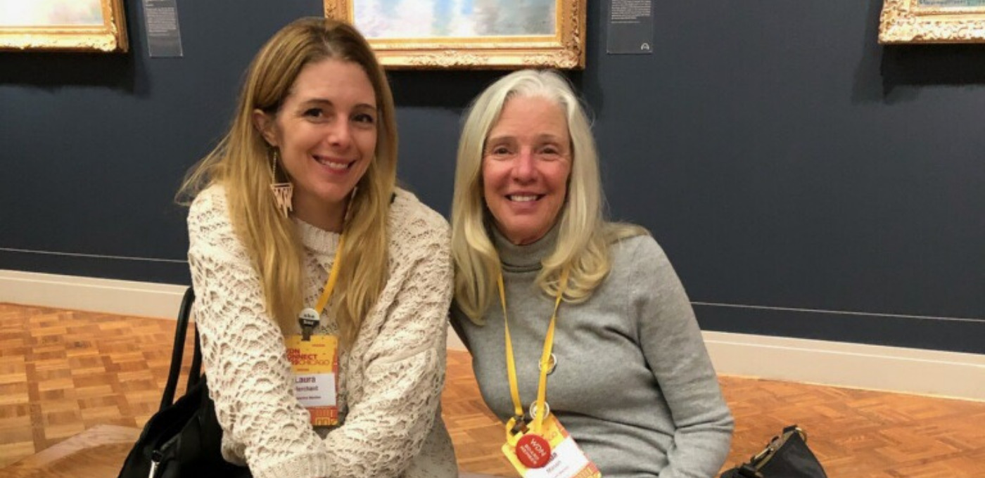 A photo of Linda Mason and Laura Merchant. They sit side by side on a bench at an art gallery and smile at the camera.