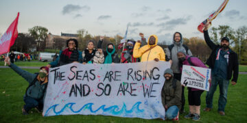 A photo of a group of people waving flags and standing behind a large banner that reads "the seas are rising and so are we"