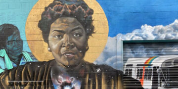 a mural of dorothy bolden. She is painted on a wall with a halo behind her head and colors over her heart. Another person and the sky is in the background
