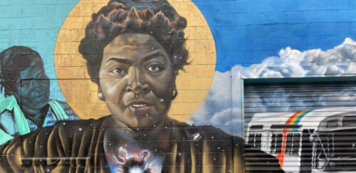 a mural of dorothy bolden. She is painted on a wall with a halo behind her head and colors over her heart. Another person and the sky is in the background