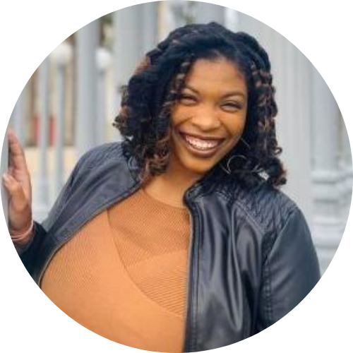 Photo of Venneikia Williams, a Black person smiling at the camera wearing a brown shirt and Black jacket with shoulder length black and brown toned hair