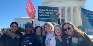 Photo of several people standing in front of a building. They hold signs that say, among other things, "I love someone who had an abortion. #MyRightMyDecision"