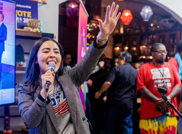 Photo of a person in a blazer and a shirt that says "vote" speaking into a microphone and raising up their hand