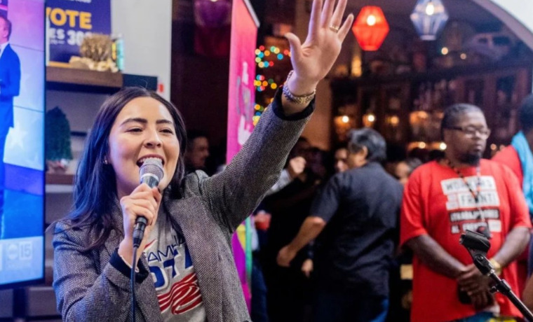 Photo of a person in a blazer and a shirt that says "vote" speaking into a microphone and raising up their hand